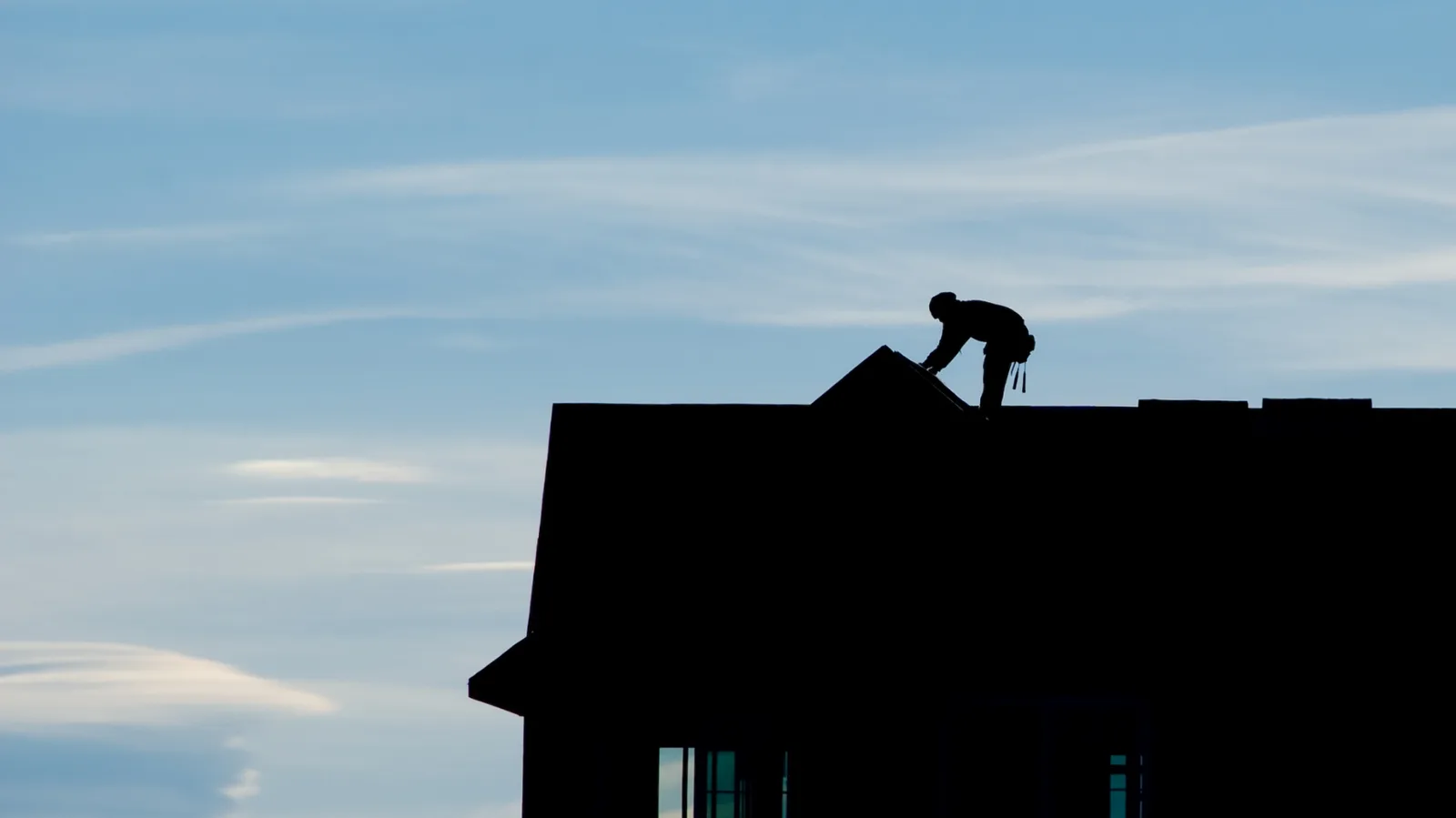 Distant backlit roofer working along the roofline of a house